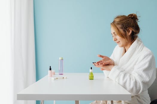 Woman in bathrobe applying skincare products at a table in a bright studio setting.